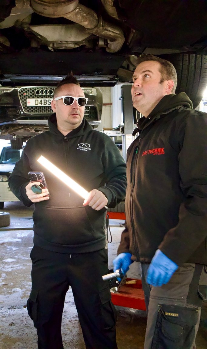White Shades Media filming in a garage while a mechanic discusses work under a raised car