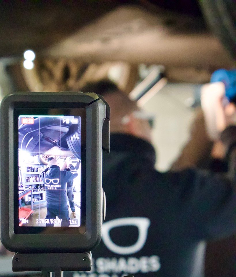 Action camera screen showing White Shades Media filming a mechanic working under a vehicle in a garage